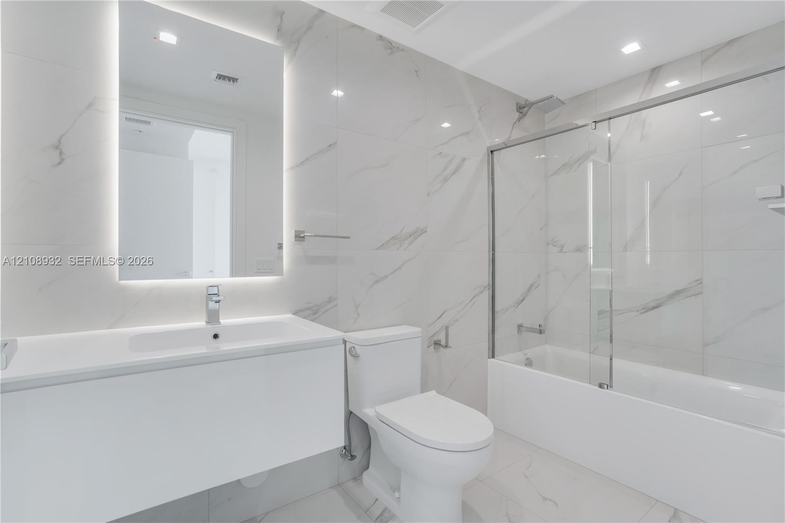 Bathroom featuring large-format stone-patterned wall and floor tile, a modern floating vanity with an integrated sink, a frameless glass tub enclosure, and an LED-backlit mirror