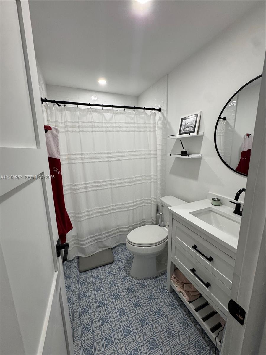 Bathroom featuring patterned tile flooring, white vanity with integrated sink and matte black hardware, round vanity mirror, recessed ceiling lighting, and wall-mounted shelving