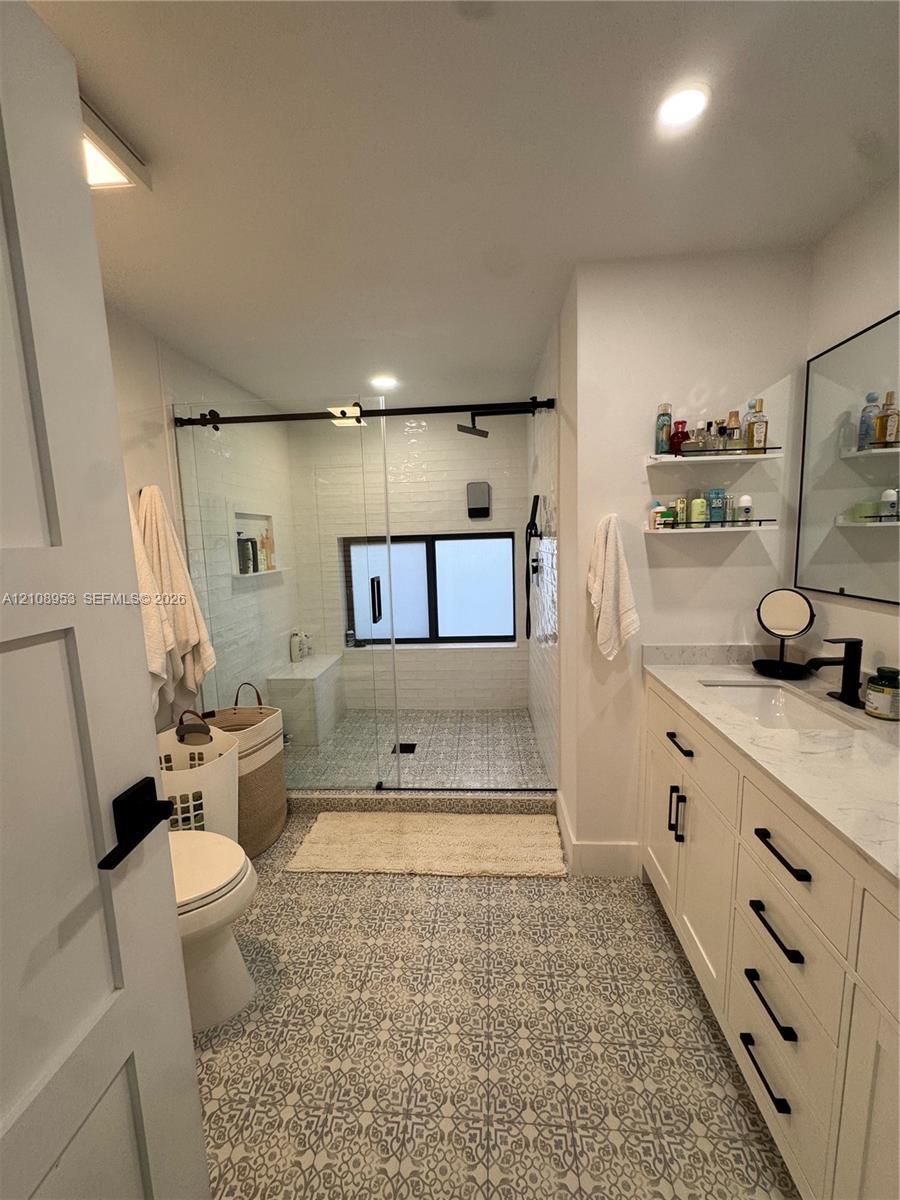 Custom cabinetry with dark hardware, stone-top vanity, walk-in glass-enclosed shower with matte black fixtures, patterned tile flooring, and integrated wall shelving