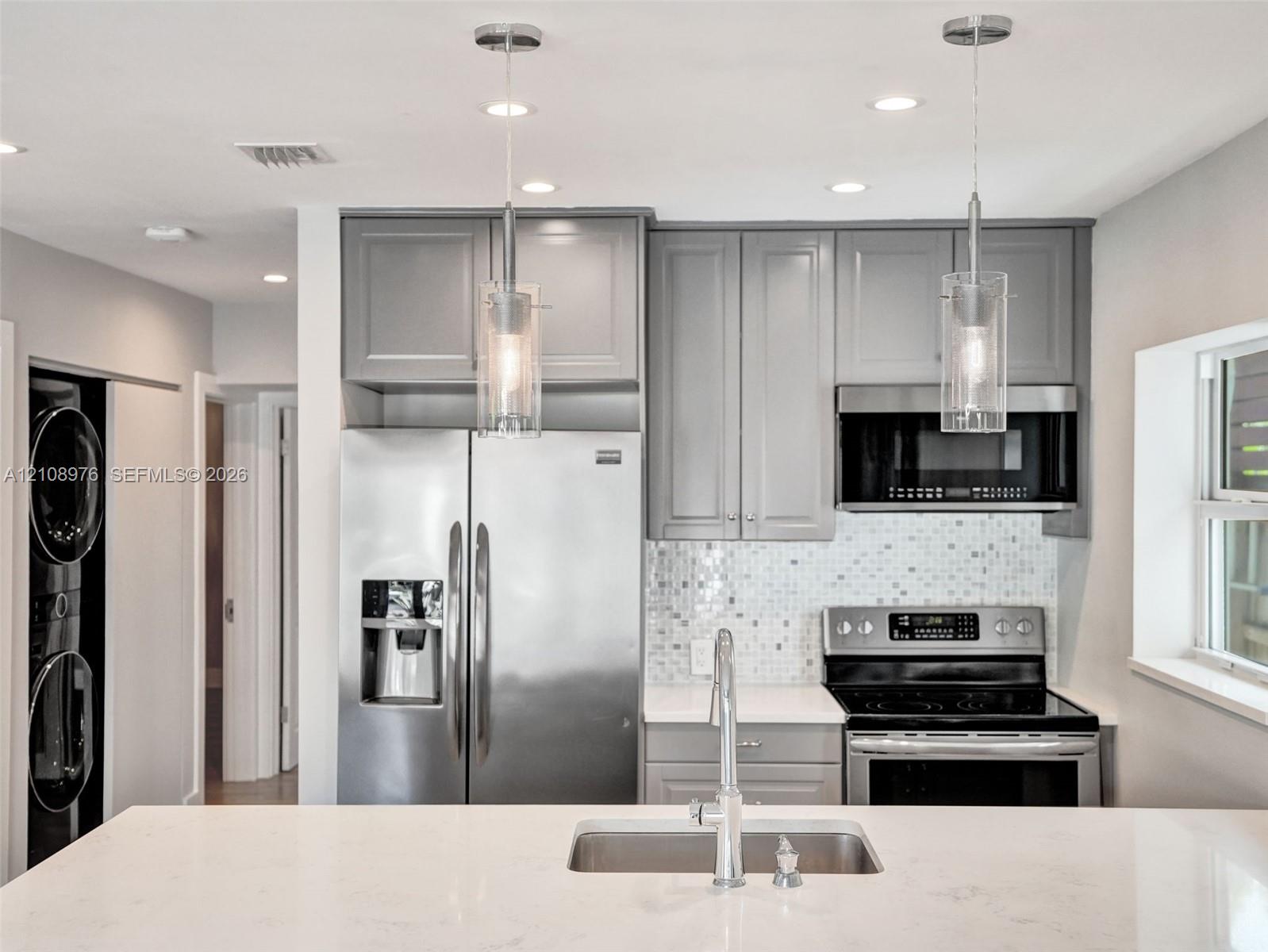 Kitchen featuring grey shaker-style cabinetry, stainless steel appliances, white stone-finish countertops, a mosaic tile backsplash, and dual glass pendant lights