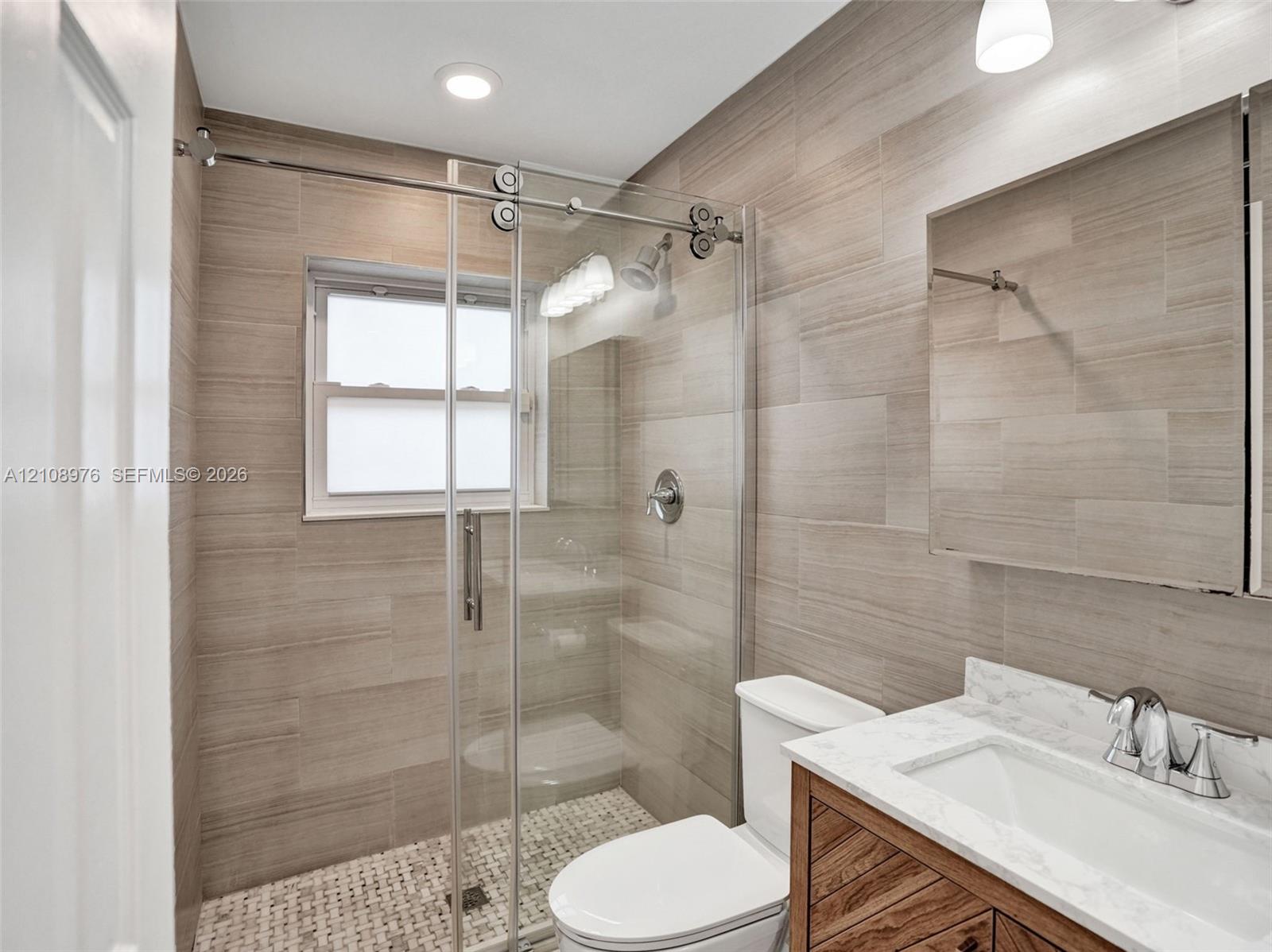 Walk-in shower with sliding glass door, mosaic floor tiling, full-height wall tile, wood-finish vanity with stone countertop, and recessed ceiling lighting