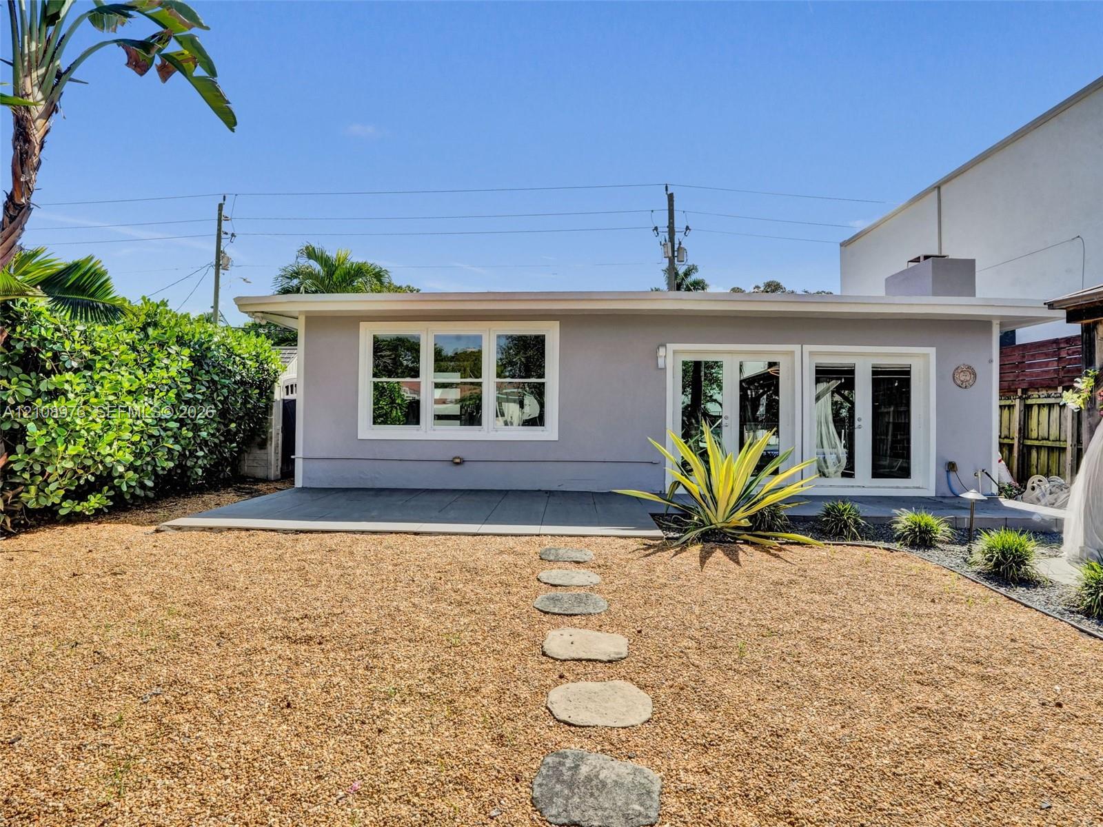 Rear exterior featuring a concrete patio, dual glass-paneled double doors, horizontal multi-pane windows, and a stone-paver walkway set within a gravel yard