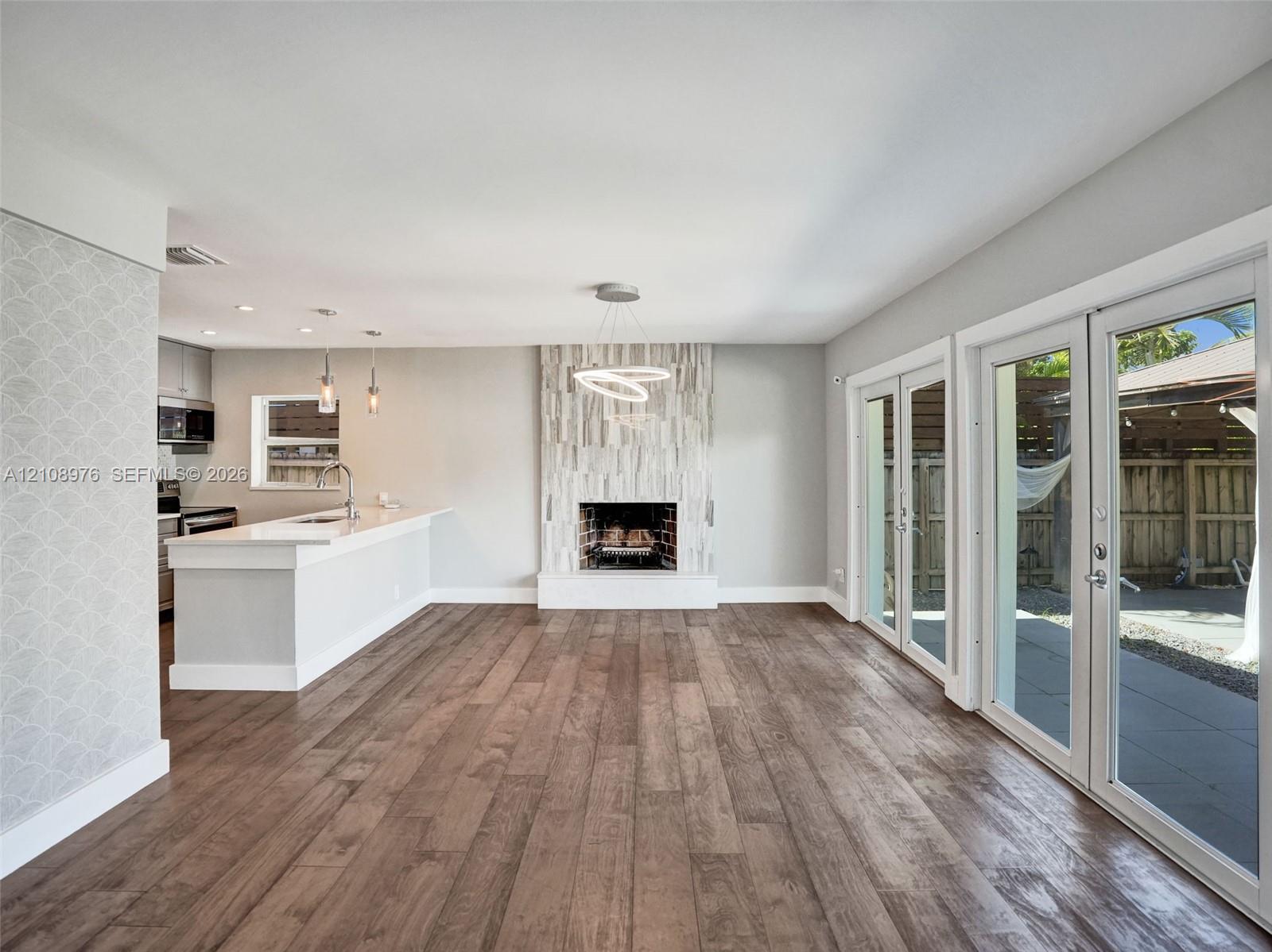 Open-concept interior featuring wood-finish flooring, floor-to-ceiling fireplace with vertical stone-finish surround, kitchen island with solid-surface countertop, dual glass-paned doors, and contemporary tiered chandelier