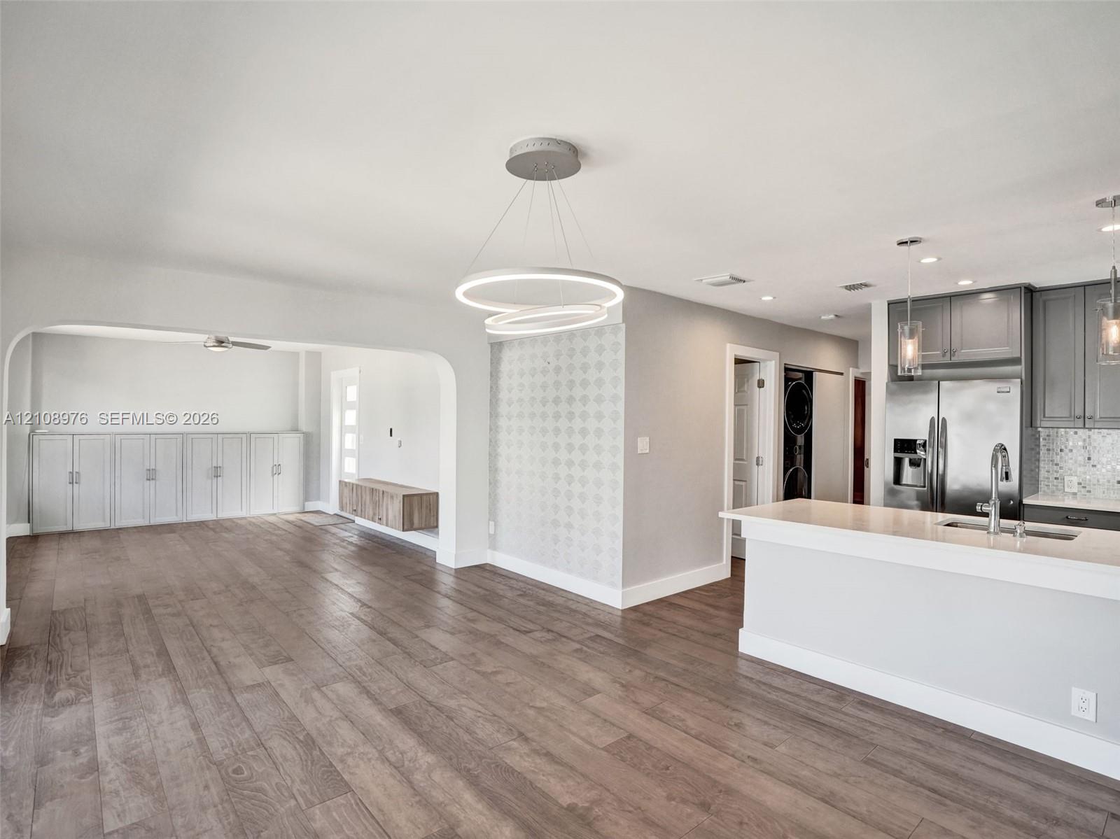 Open-concept interior featuring wood-finish flooring, a modern dual-ring pendant light fixture, cabinetry with stone countertops, and a decorative tile accent wall