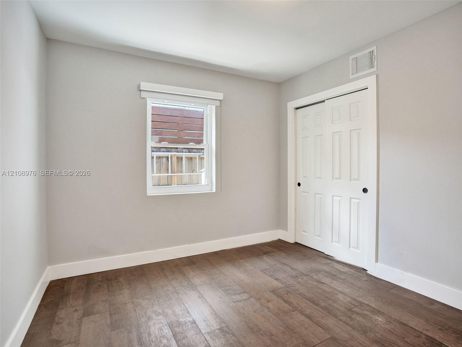 Bedroom 2. Neutral interior featuring wood-finish flooring, white paneled sliding closet doors, and a single-hung window with a classic valance