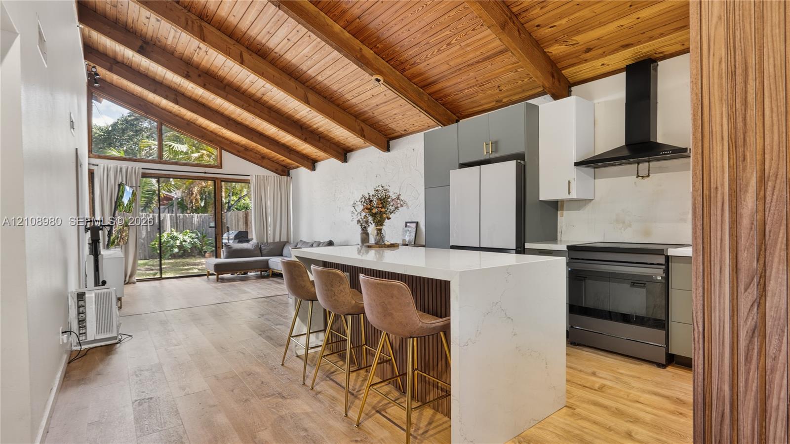 Vaulted wood-plank ceiling with exposed beams, kitchen island with a waterfall-edge countertop, wood-finish flooring, two-tone cabinetry, and matte black range hood