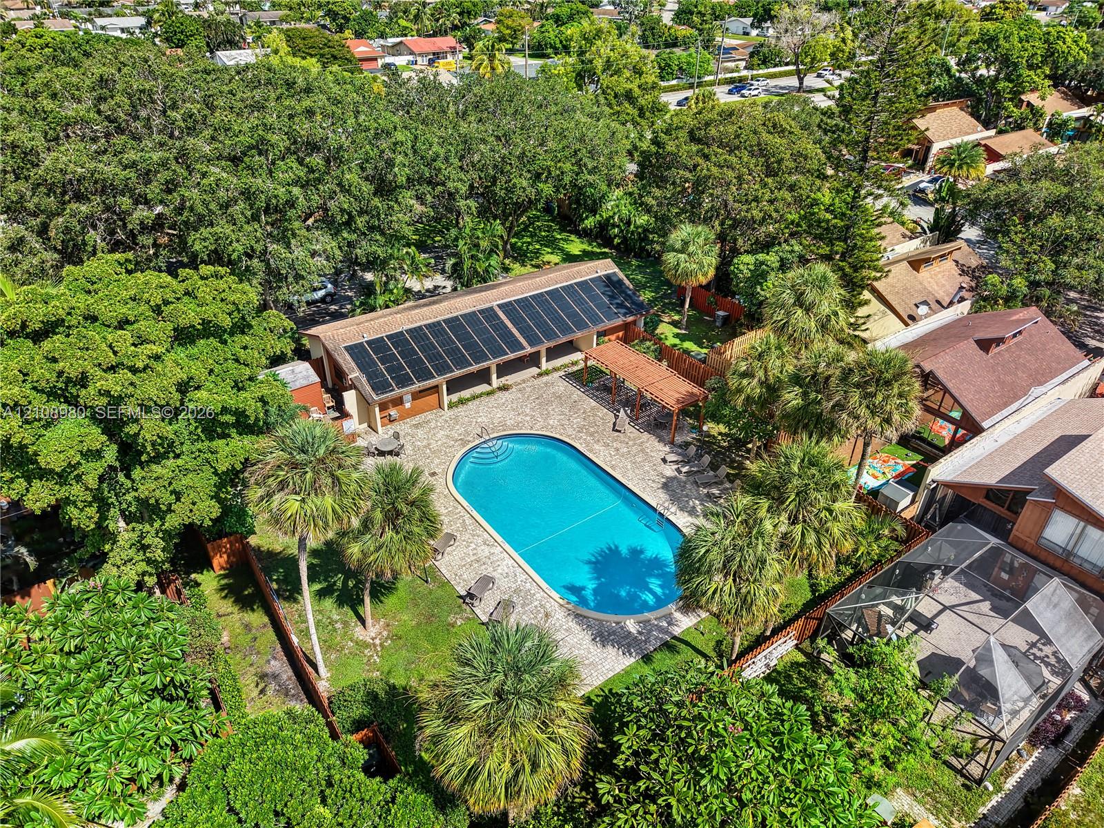 In-ground pool with surrounding paved patio, detached building with integrated solar panel array, and wood-framed pergola set within a mature tree canopy