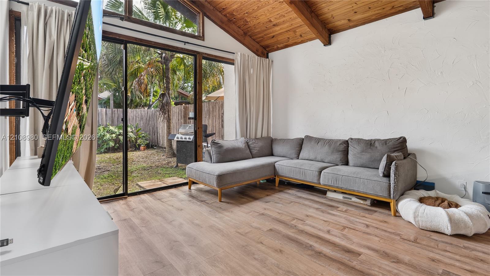 Vaulted ceiling with exposed wood beams, wood-finish flooring, textured wall finish, and floor-to-ceiling sliding glass doors opening to a fenced outdoor area
