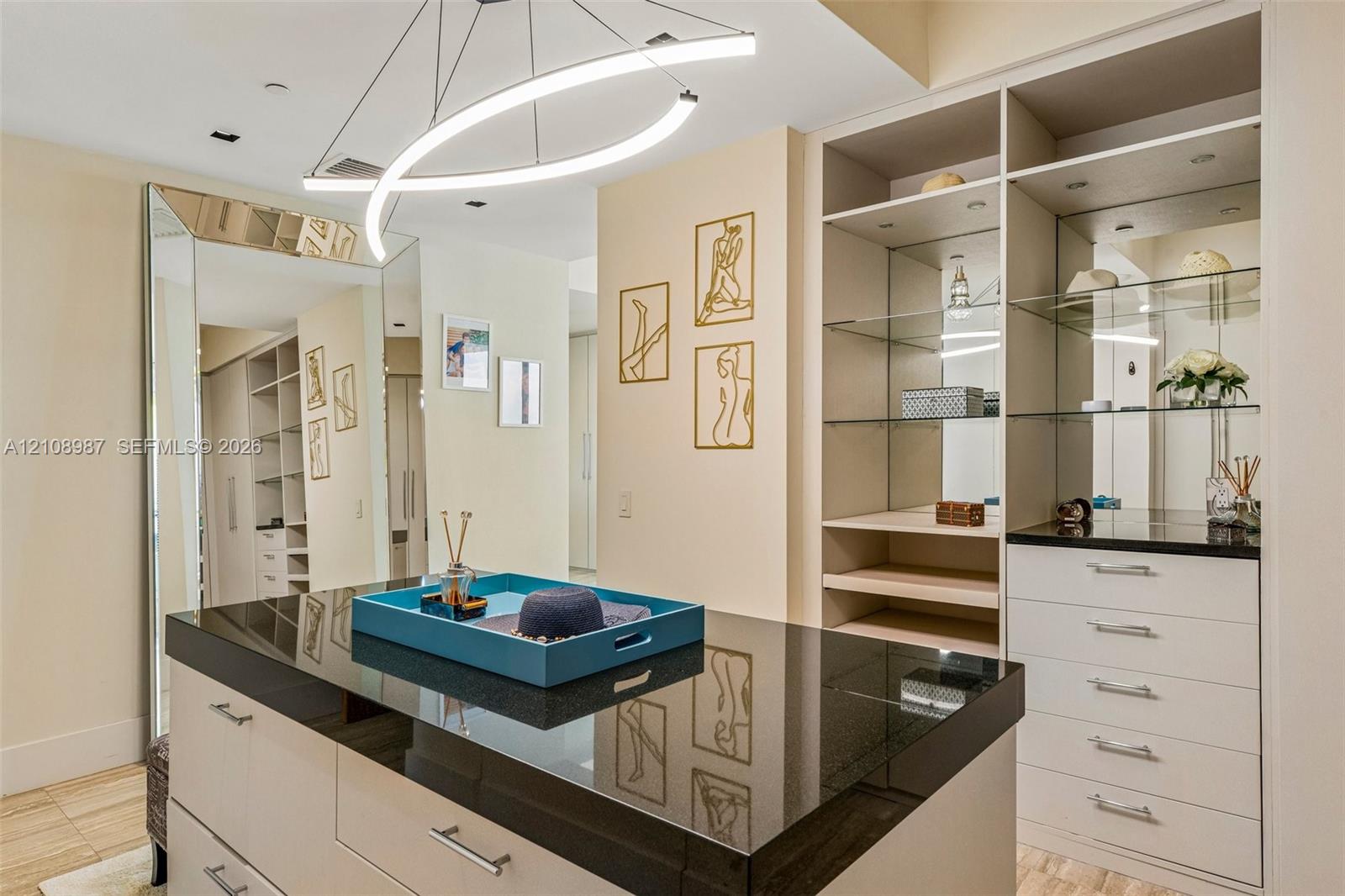 Custom walk-in closet featuring floor-to-ceiling cabinetry, a central storage island with a dark stone countertop, built-in glass shelving, mirrored paneling, and a contemporary curved light fixture