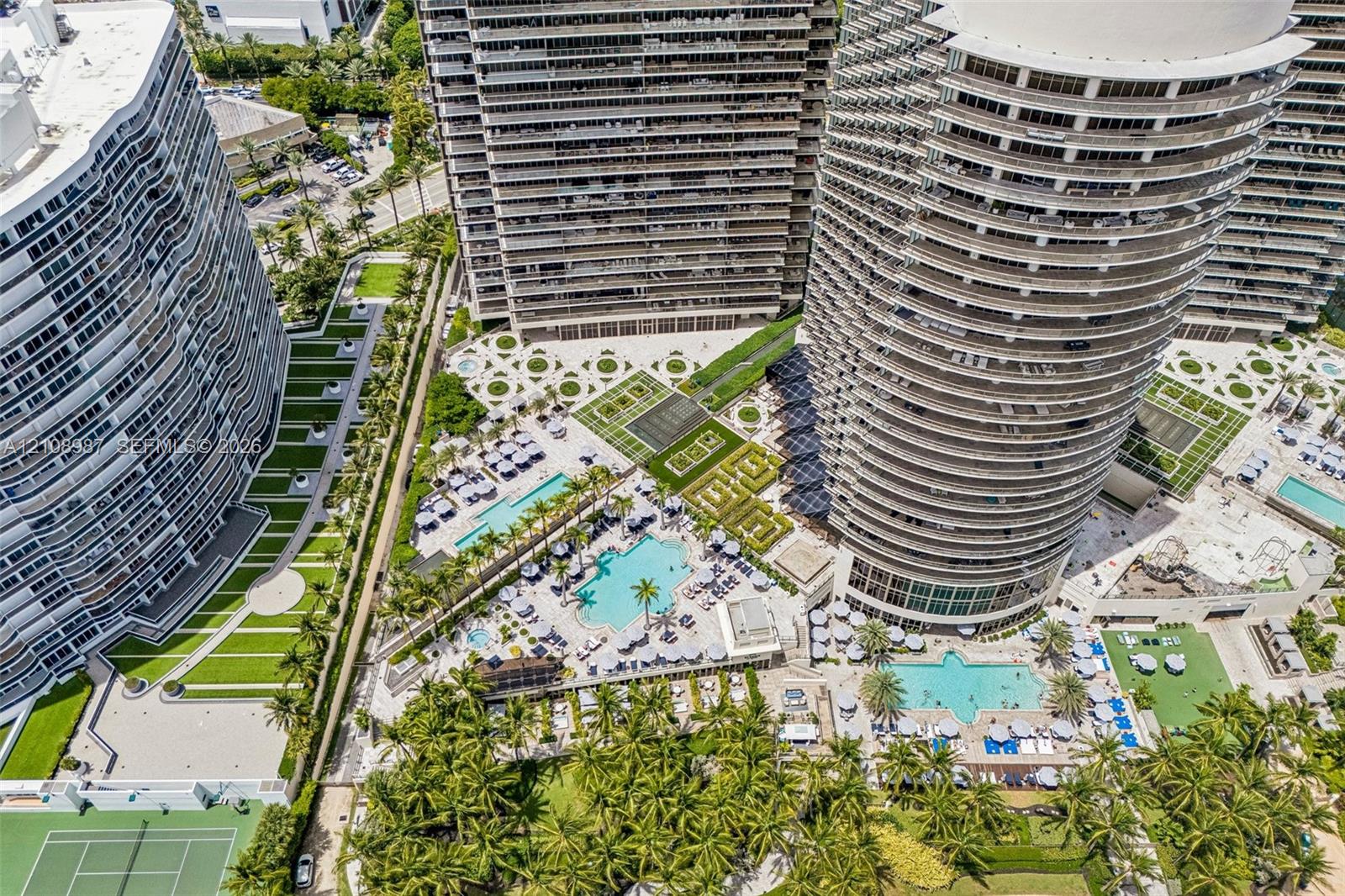 Aerial perspective of a high-rise residential complex featuring multiple swimming pools, manicured terrace gardens, paved courtyards, and a tennis court