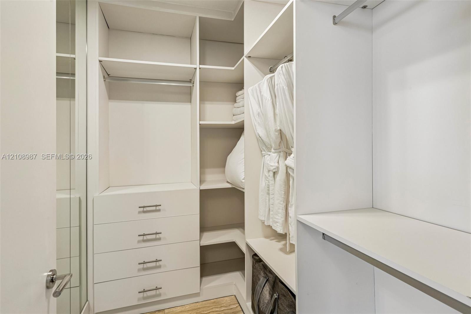 Custom walk-in closet featuring white built-in cabinetry, floor-to-ceiling shelving, integrated hanging rods, and a four-drawer dresser unit with metallic pulls