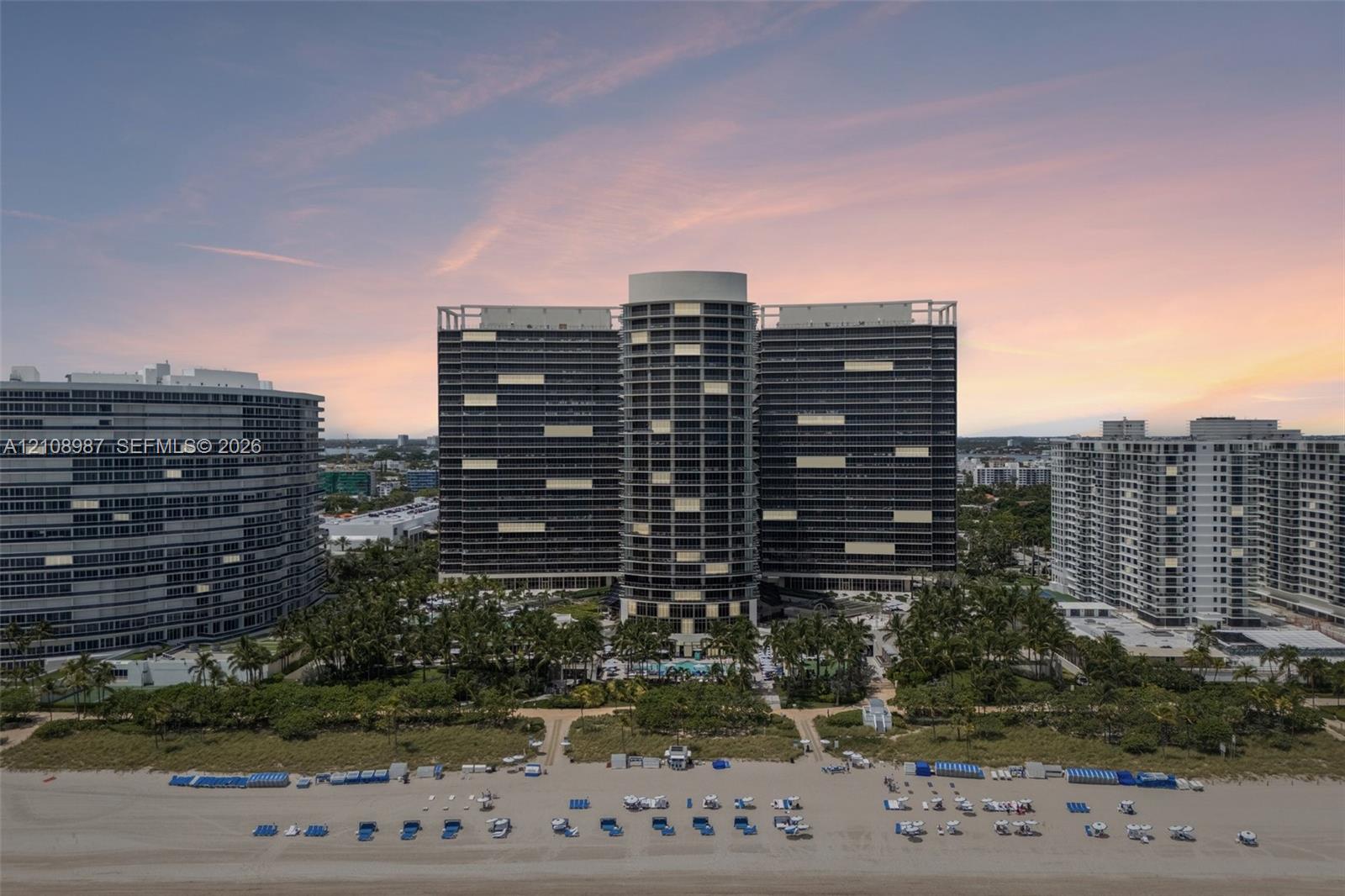 High-rise beachfront structure featuring wraparound balconies, expansive glass facade, landscaped grounds with mature palm trees, and direct sand access