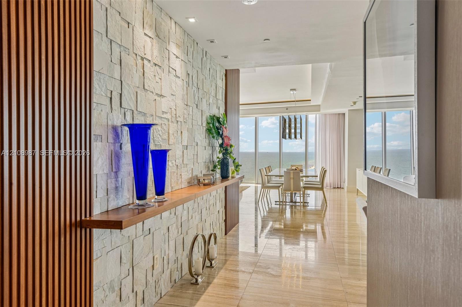 Textured stone accent wall with a wood-finished floating shelf, vertical wood-slat paneling, polished stone flooring, and floor-to-ceiling windows