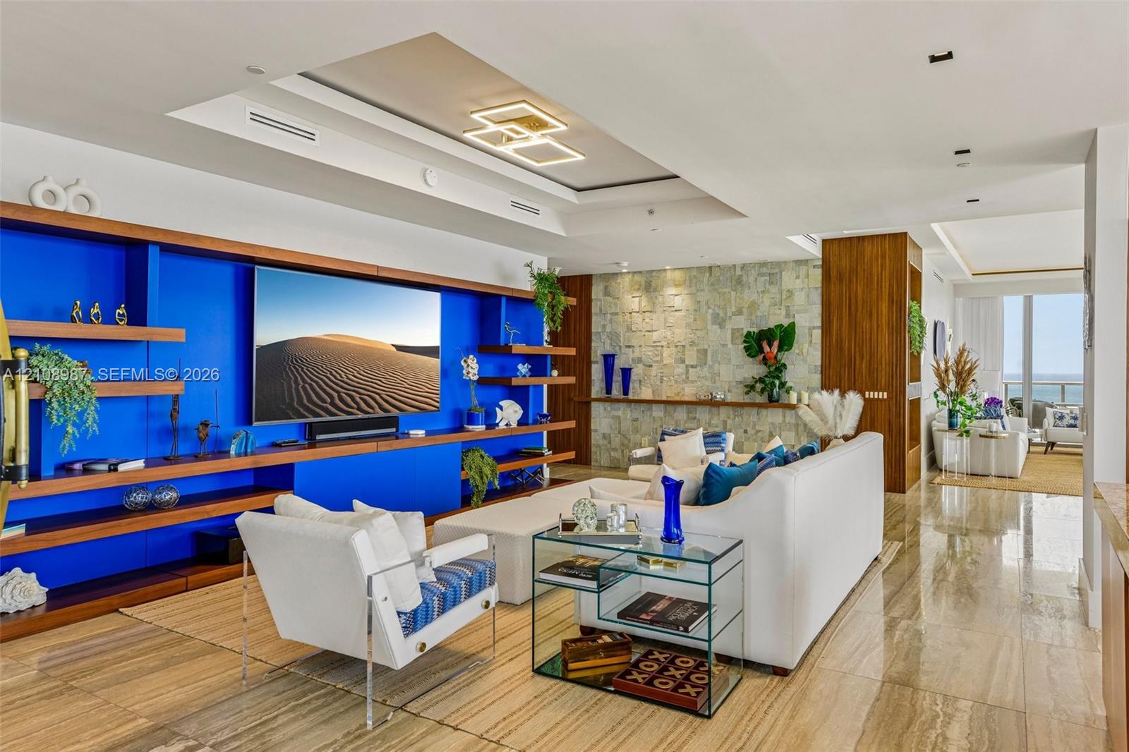 Living area featuring large-format stone tile flooring, custom wood-finish built-in shelving, a natural stone accent wall, recessed tray ceiling, and geometric integrated ceiling lighting