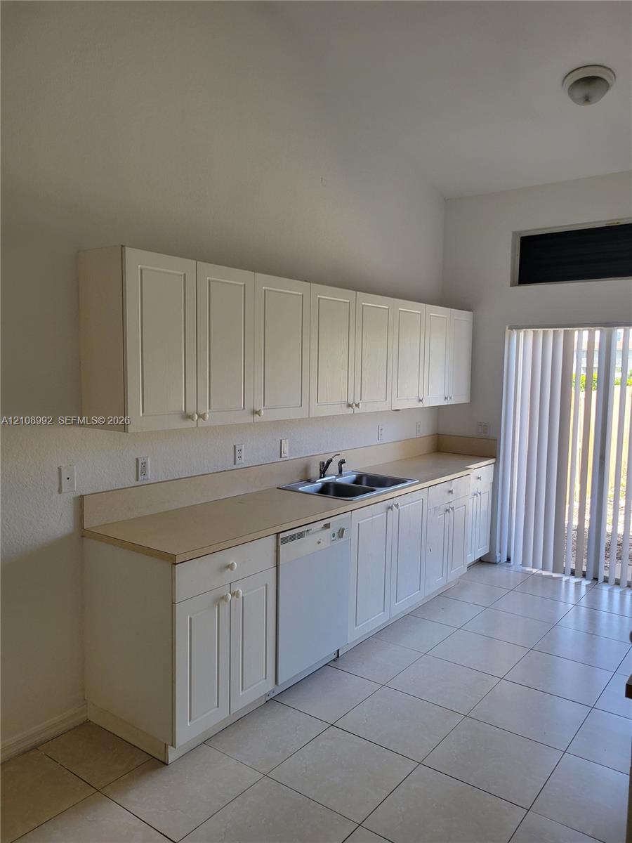 Kitchen featuring white shaker-style cabinetry, solid-surface countertops, dual-basin stainless steel sink, integrated dishwasher, and tile flooring