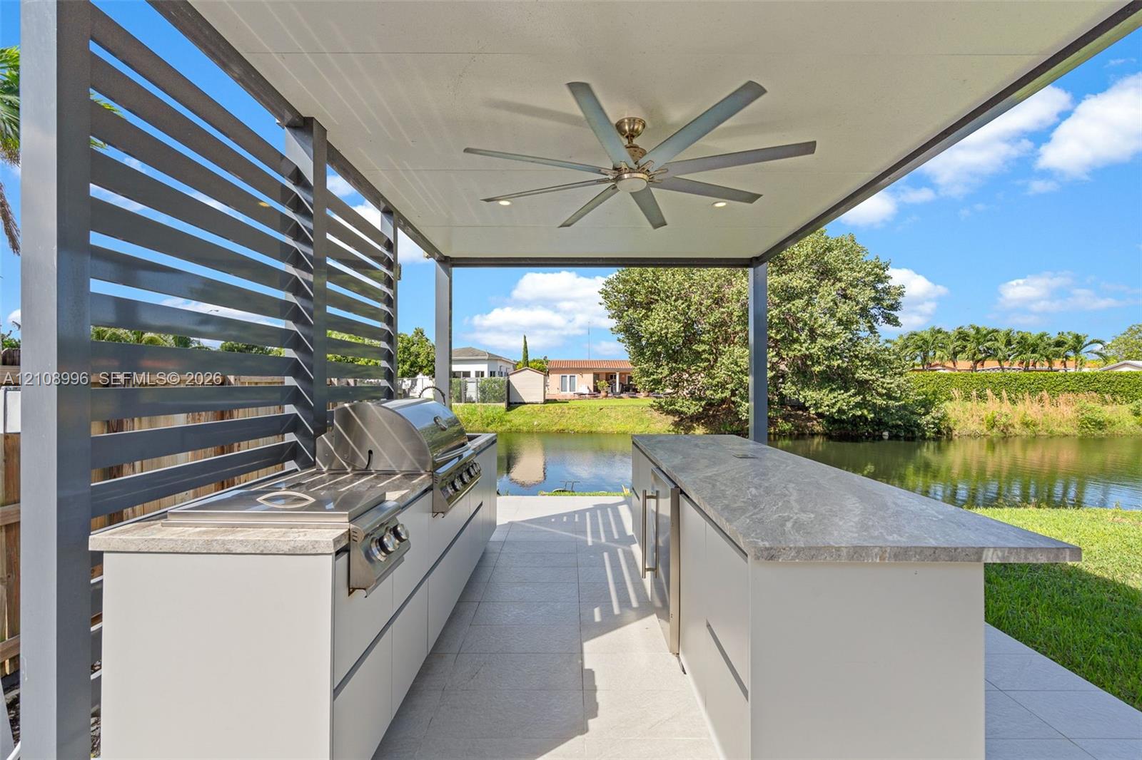 Covered outdoor kitchen featuring a stone-topped island, stainless steel grill, horizontal slat privacy screen, and an integrated ceiling fan