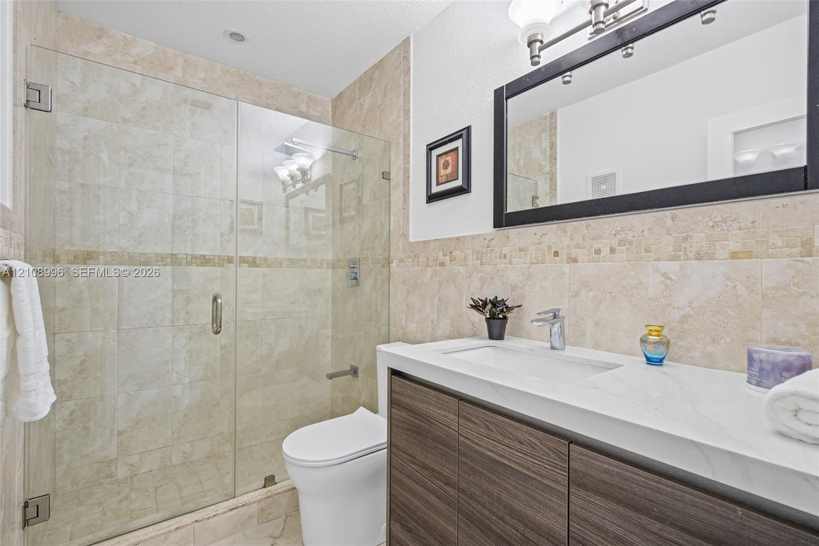 Bathroom featuring a vanity with wood-finish cabinetry, a white countertop with an integrated sink, a frameless glass-enclosed shower with neutral tile surround, and a metal-framed mirror
