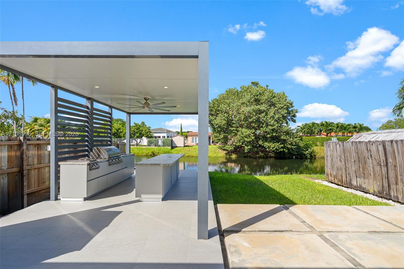 Outdoor kitchen featuring a built-in stainless steel grill, stone-topped island, modern pergola structure with recessed lighting and a ceiling fan, and large-format paved patio flooring