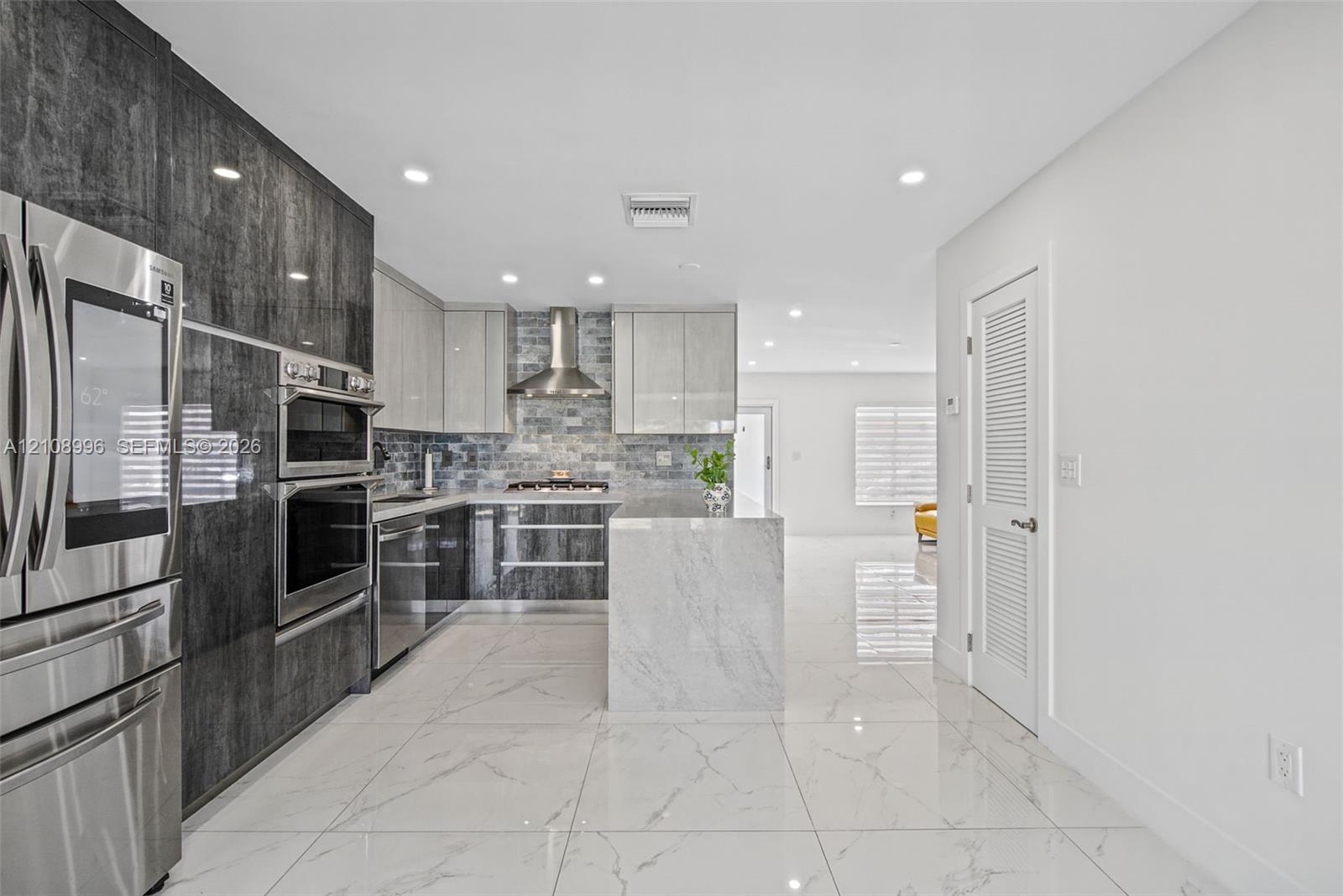 Kitchen featuring two-tone cabinetry with high-gloss finishes, a center island with stone-finish waterfall edges, stainless steel built-in appliances, a glass tile backsplash, and large-format porcelain tile flooring