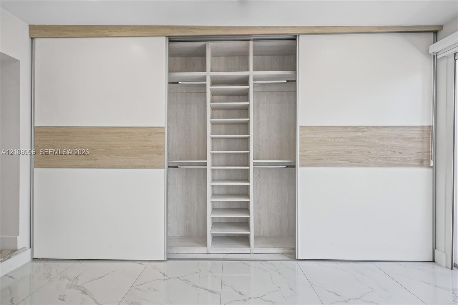 Built-in floor-to-ceiling wardrobe featuring sliding dual-tone panel doors, integrated shelving, hanging rods, and light wood-finish interior detailing, complemented by large-format polished tile flooring