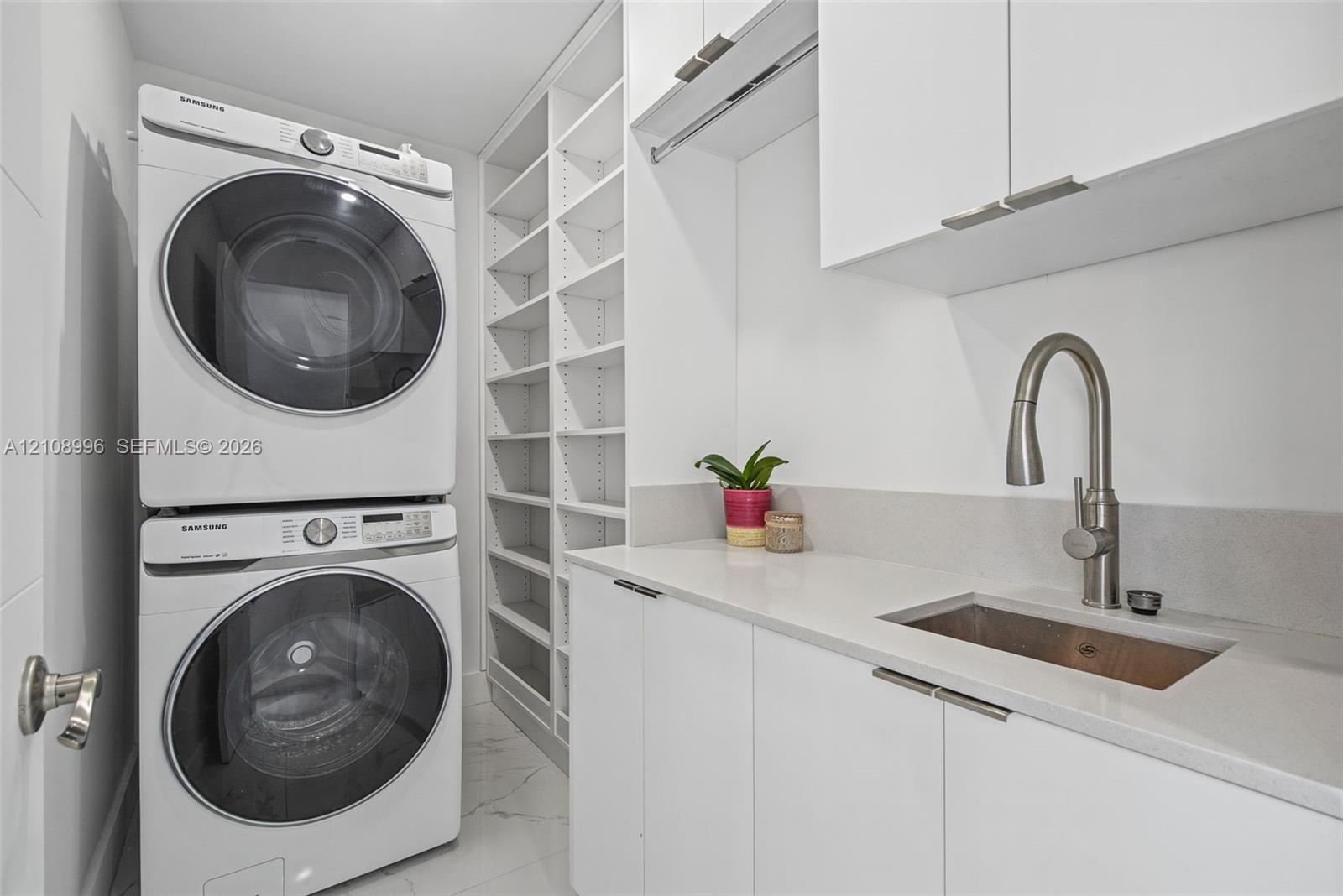 Laundry area featuring custom white cabinetry, stone-surface countertop with an integrated basin, high-arc pull-down faucet, floor-to-ceiling built-in shelving, and large-format tile flooring