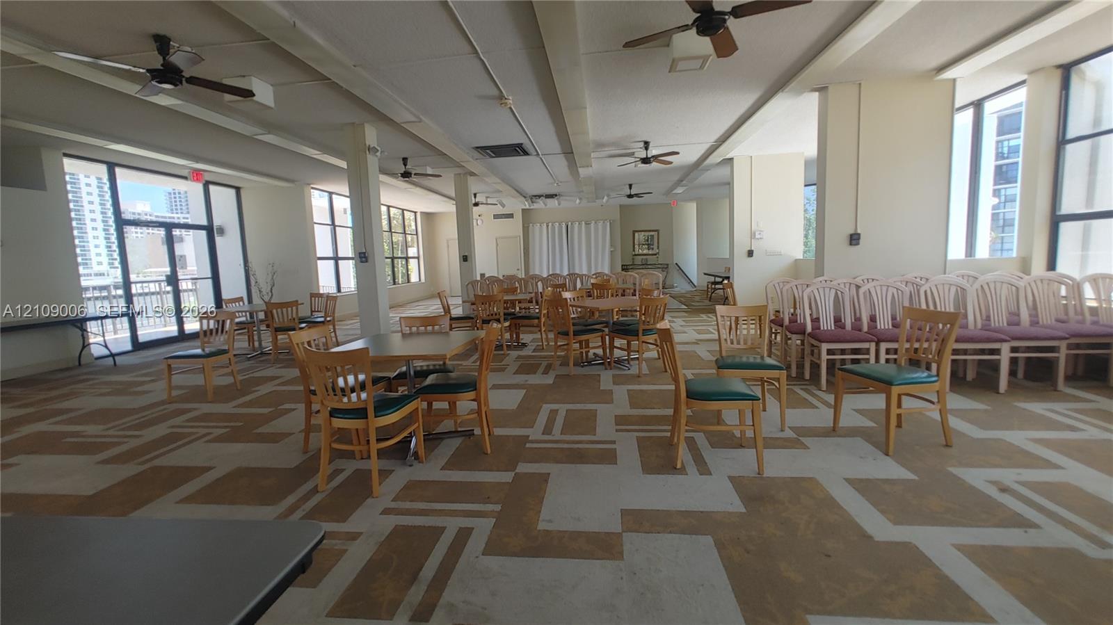 Expansive interior space featuring patterned carpeting, multiple ceiling fans, floor-to-ceiling window panels, and neutral wall tones
