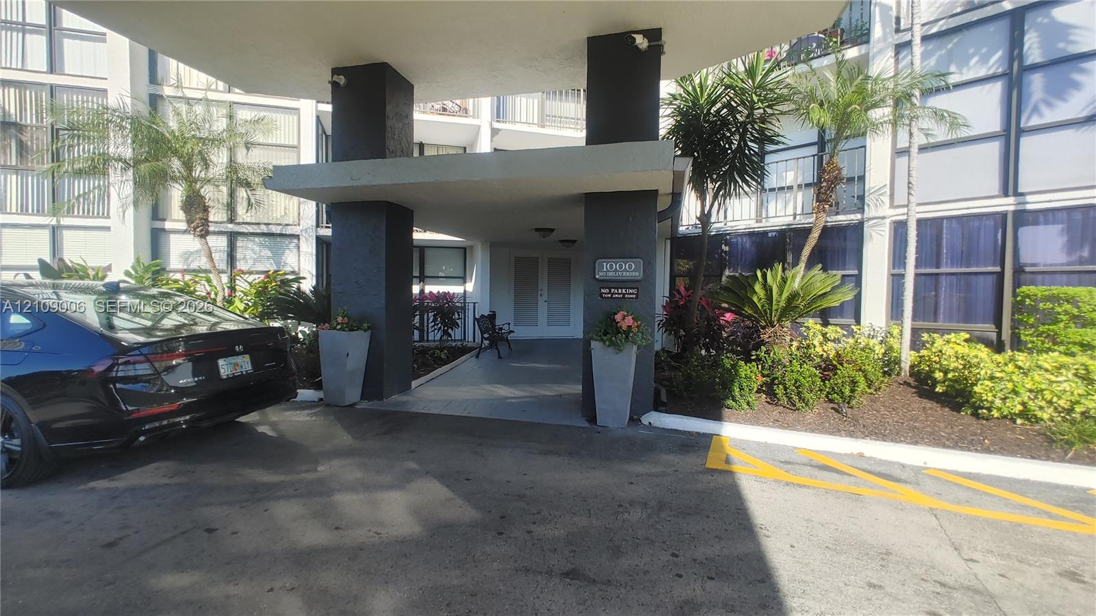 Covered building entrance featuring a concrete porte-cochre, decorative structural pillars, and curated landscape planters