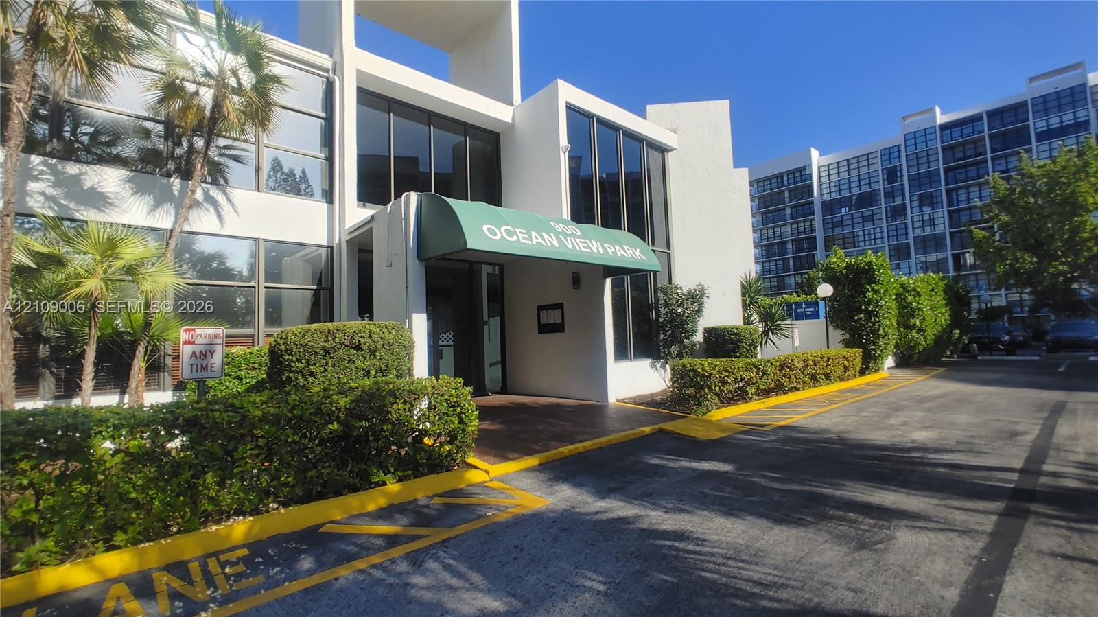 Multi-story building exterior featuring a white stucco facade, expansive floor-to-ceiling glass windows, and mature perimeter landscaping