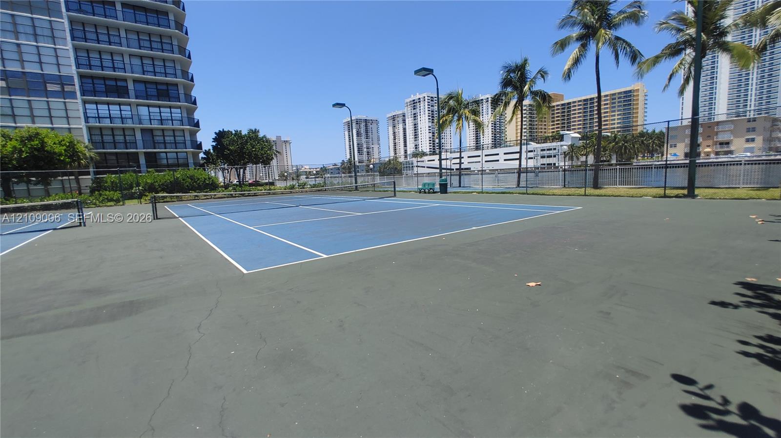Hard-surface tennis court featuring perimeter fencing, integrated lighting, and palm tree landscaping