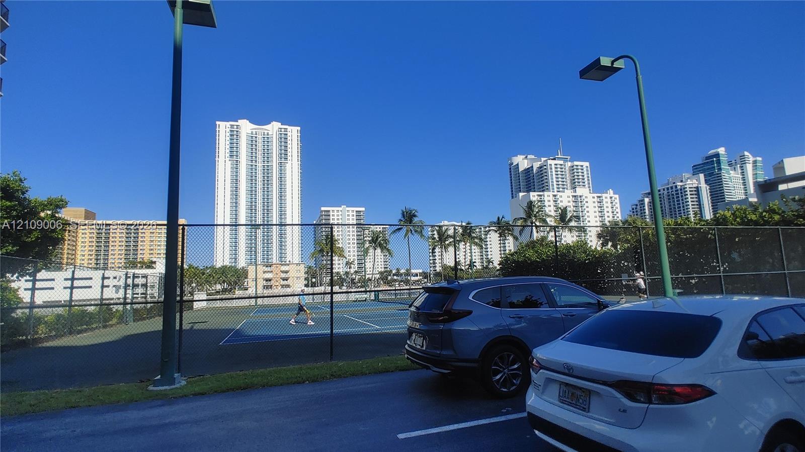 Fenced tennis court with a hard surface, tall perimeter pole lighting, and asphalt parking area