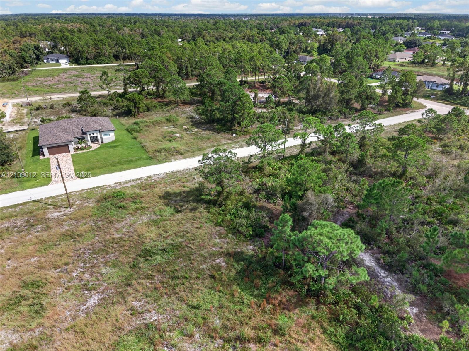 Aerial perspective of a detached residential property featuring a paver driveway, an attached garage, a multi-textured exterior facade, and expansive surrounding grounds with mature pine trees and native vegetation