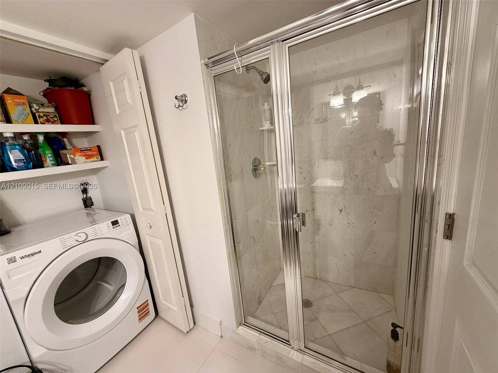 Walk-in shower enclosure with glass doors, stone-finish wall tiling, built-in shelving, chrome-finish plumbing fixtures, and integrated laundry area with overhead storage shelving