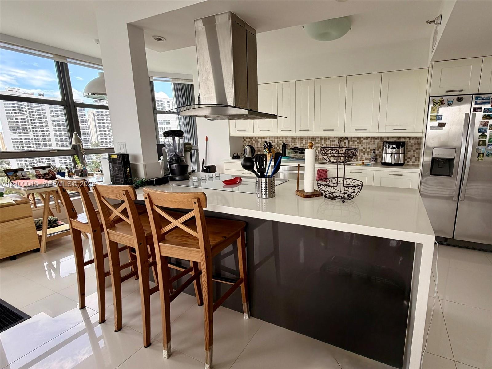 Kitchen featuring an island with a solid-surface countertop, stainless steel chimney-style range hood, white shaker-style cabinetry, mosaic tile backsplash, and floor-to-ceiling windows Kitchen featuring an island with a solid-surface countertop, stainless steel chimney-style range hood, white shaker-style cabinetry, mosaic tile backsplash, and floor-to-ceiling windows