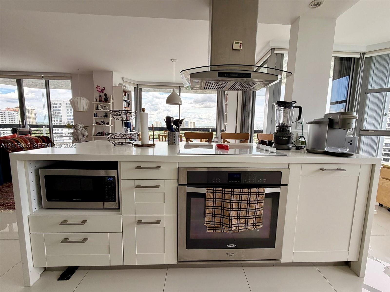Kitchen island featuring white cabinetry, a light-toned solid surface countertop, stainless steel oven, integrated microwave, and an overhead stainless steel range hood Kitchen island featuring white cabinetry, a light-toned solid surface countertop, stainless steel oven, integrated microwave, and an overhead stainless steel range hood