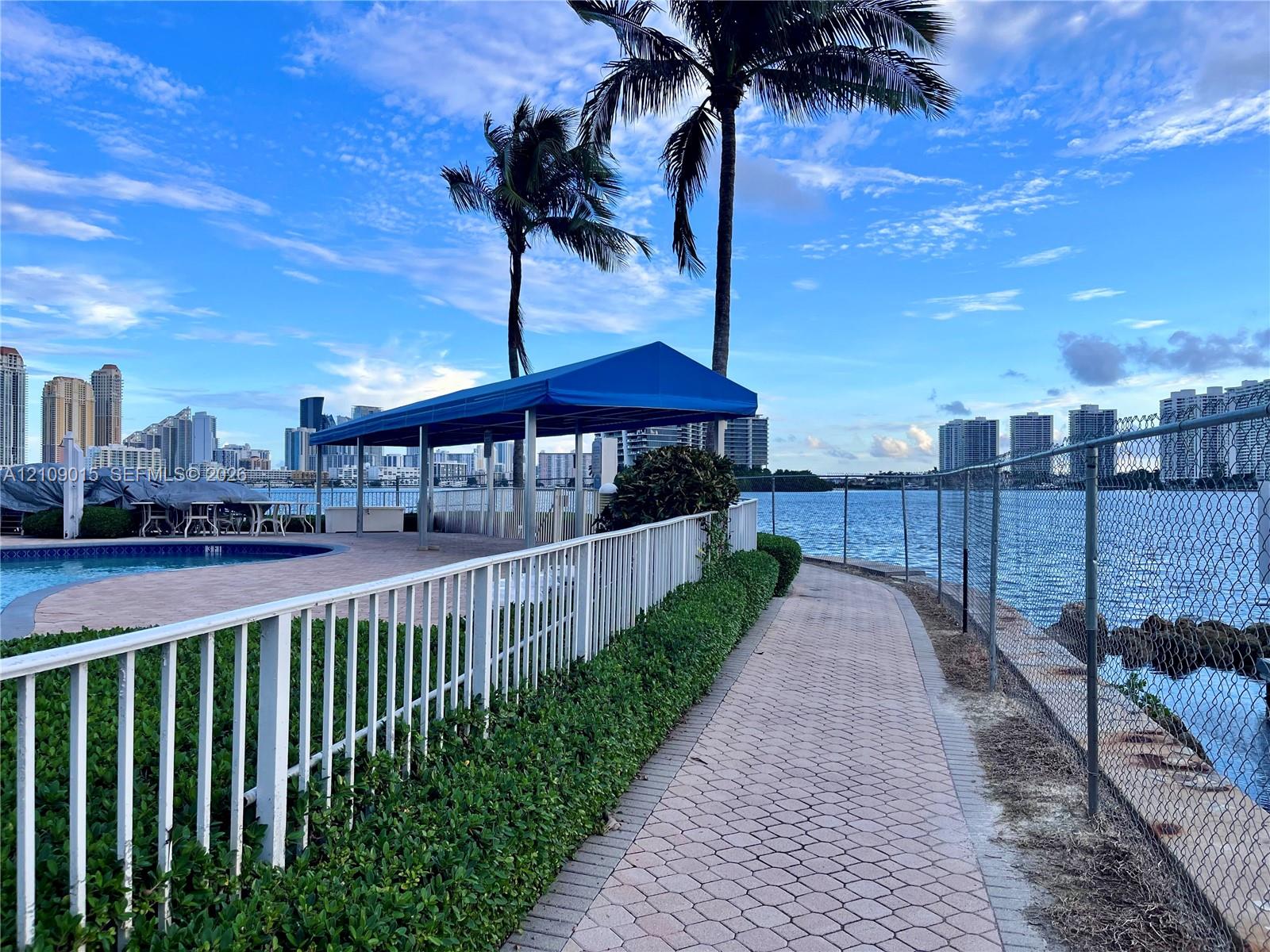 Waterfront paver walkway featuring metal perimeter fencing, mature palm trees, a poolside canopy structure, and proximity to the canal