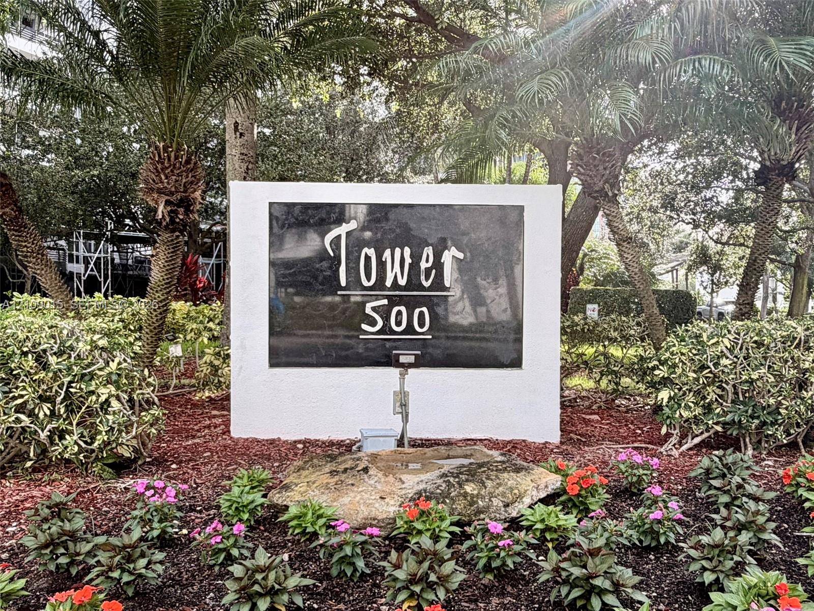 Monument signage with stone-finished display and landscaped grounds featuring mature palm trees, flowering shrubs, and decorative mulch beds