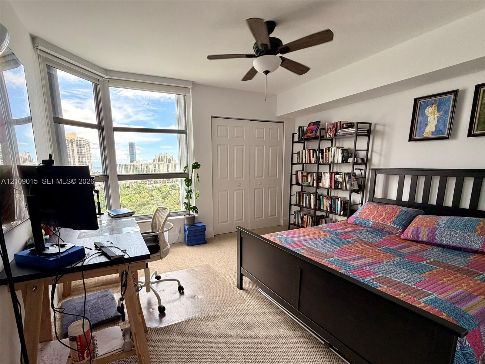 Expansive bay windows, neutral carpeting, ceiling fan with integrated lighting, and a double-door closet