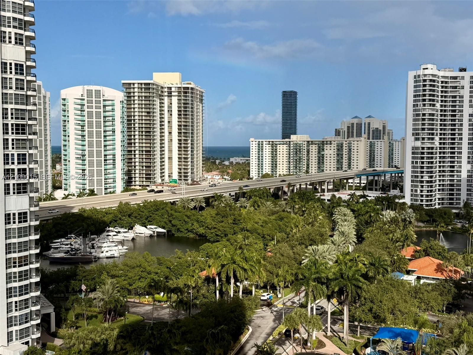 Panoramic cityscape featuring high-rise architecture, a coastal waterway with boat docks, lush palm trees, and an ocean horizon