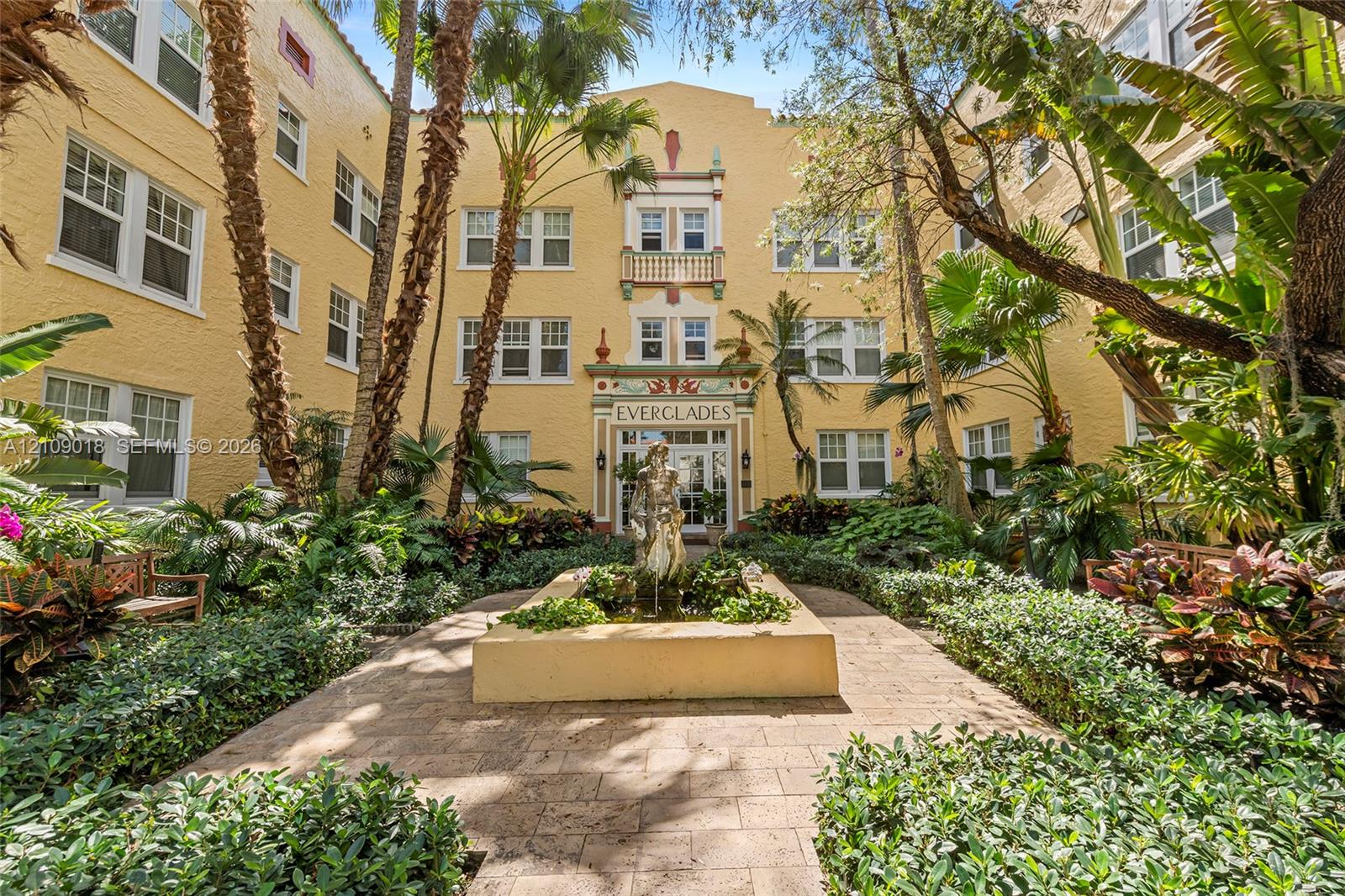 Stucco exterior featuring decorative masonry accents, a central courtyard fountain, multi-pane windows, and paved walkways surrounded by lush mature landscaping Stucco exterior featuring decorative masonry accents, a central courtyard fountain, multi-pane windows, and paved walkways surrounded by lush mature landscaping
