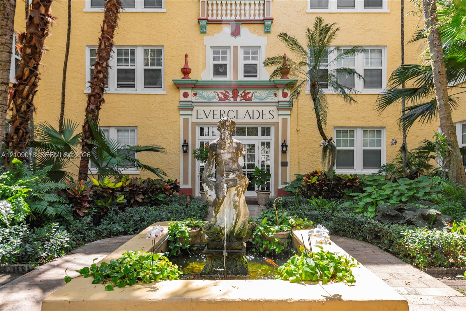 Mediterranean-style exterior featuring stucco facade, ornate architectural detailing, central fountain with sculptural centerpiece, and landscaped garden beds