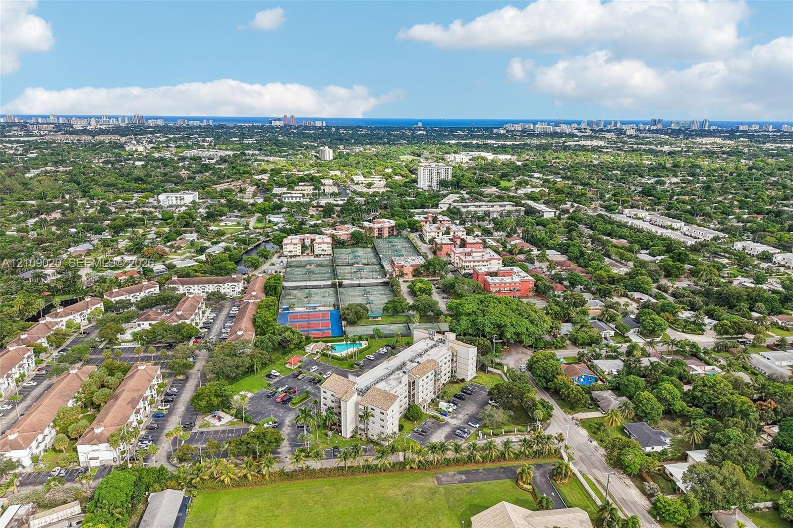 Aerial perspective of a multi-building residential complex featuring tennis courts, a swimming pool, surface parking, and landscaped grounds