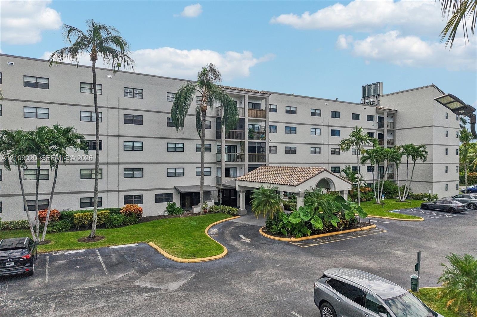 Multi-story residential building featuring a covered portico entrance with clay tile roofing, manicured green space, and palm tree landscaping