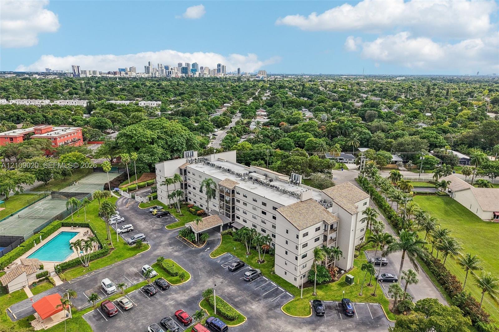 Multi-story residential complex featuring surface parking, an outdoor swimming pool, and dedicated tennis courts, set within a landscaped perimeter