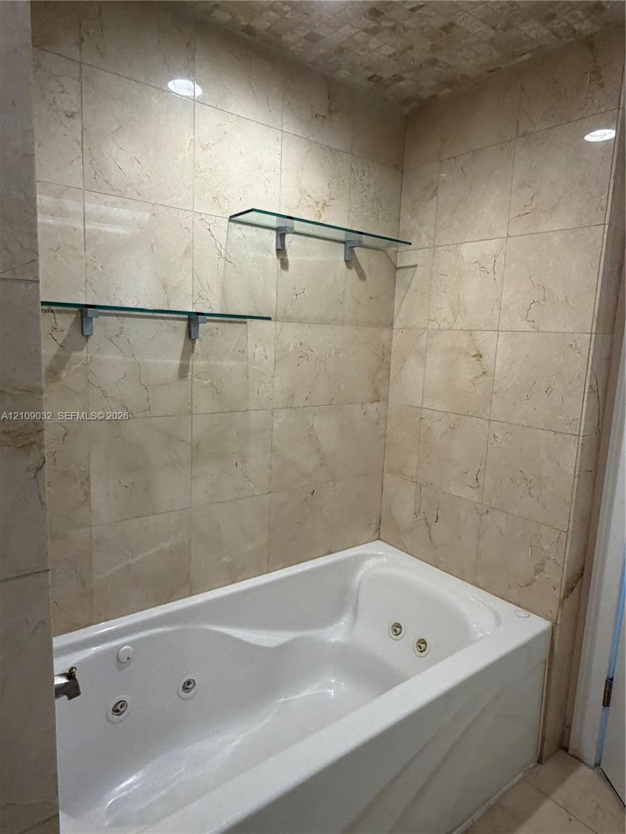 Jetted soaking tub surrounded by floor-to-ceiling stone-tile walls, complemented by dual floating glass shelves and recessed ceiling lighting