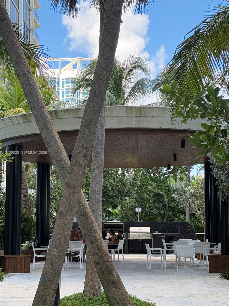 Covered outdoor pavilion featuring a wood-paneled ceiling, recessed lighting, black structural columns, and a built-in stone-finish grill station