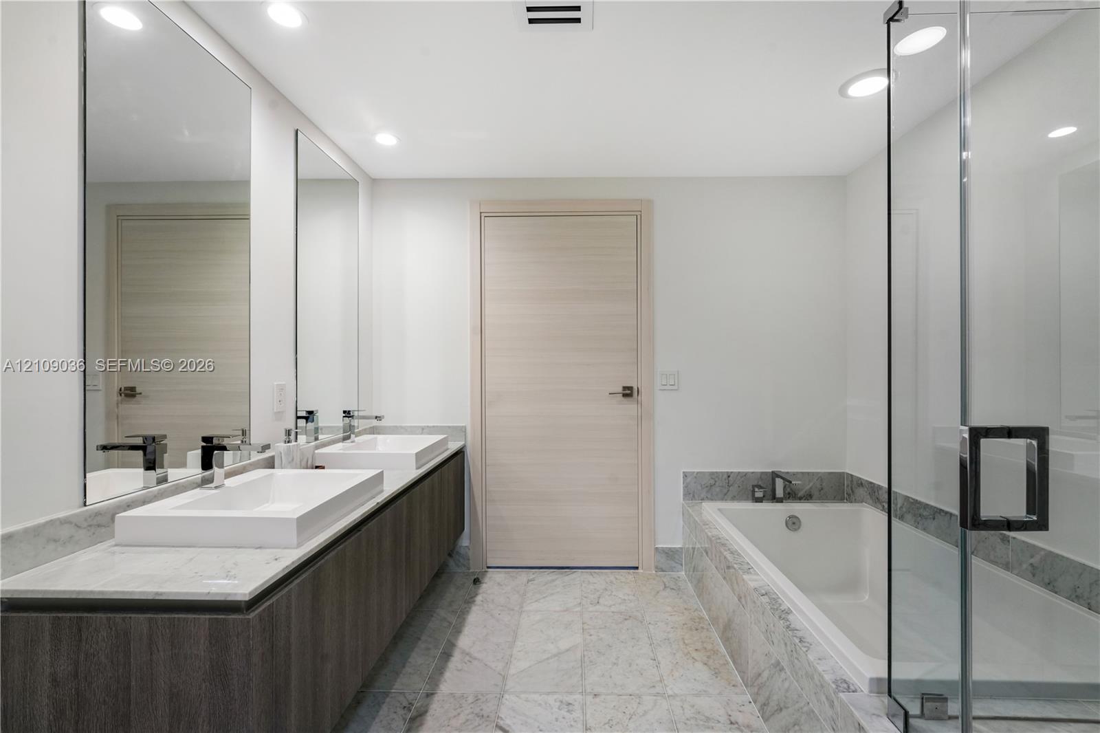 Floating double vanity with dual rectangular vessel sinks, stone-surface countertop, floor-to-ceiling mirrors, recessed lighting, and a glass-enclosed shower with a deep soaking tub