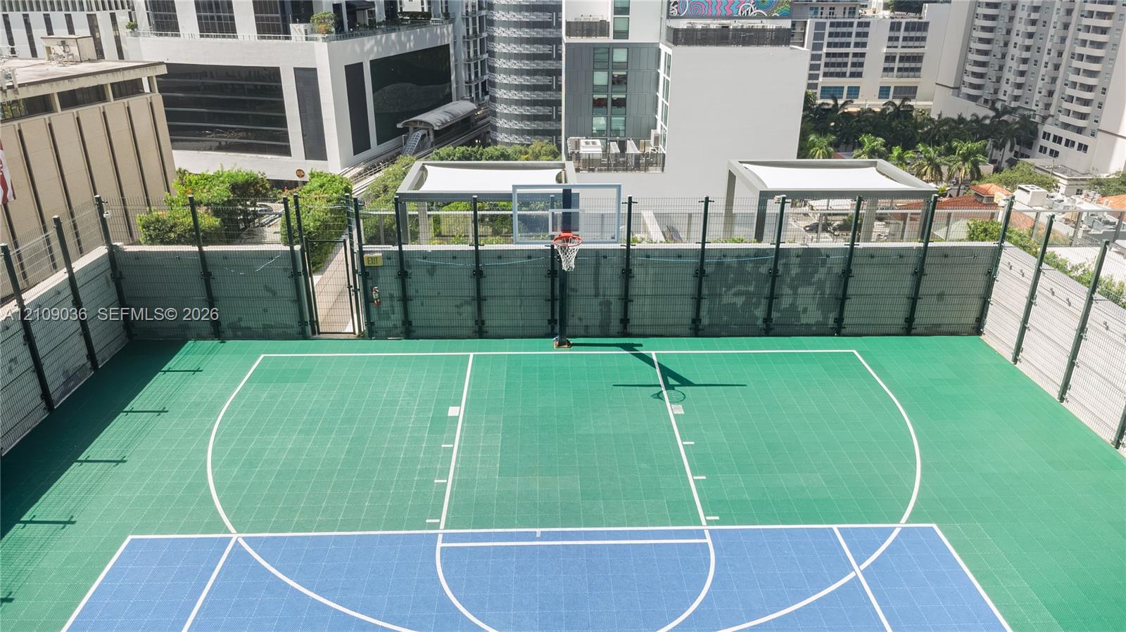 Rooftop basketball court featuring a multi-colored modular athletic surface, regulation-style hoop with glass backboard, and protective perimeter fencing