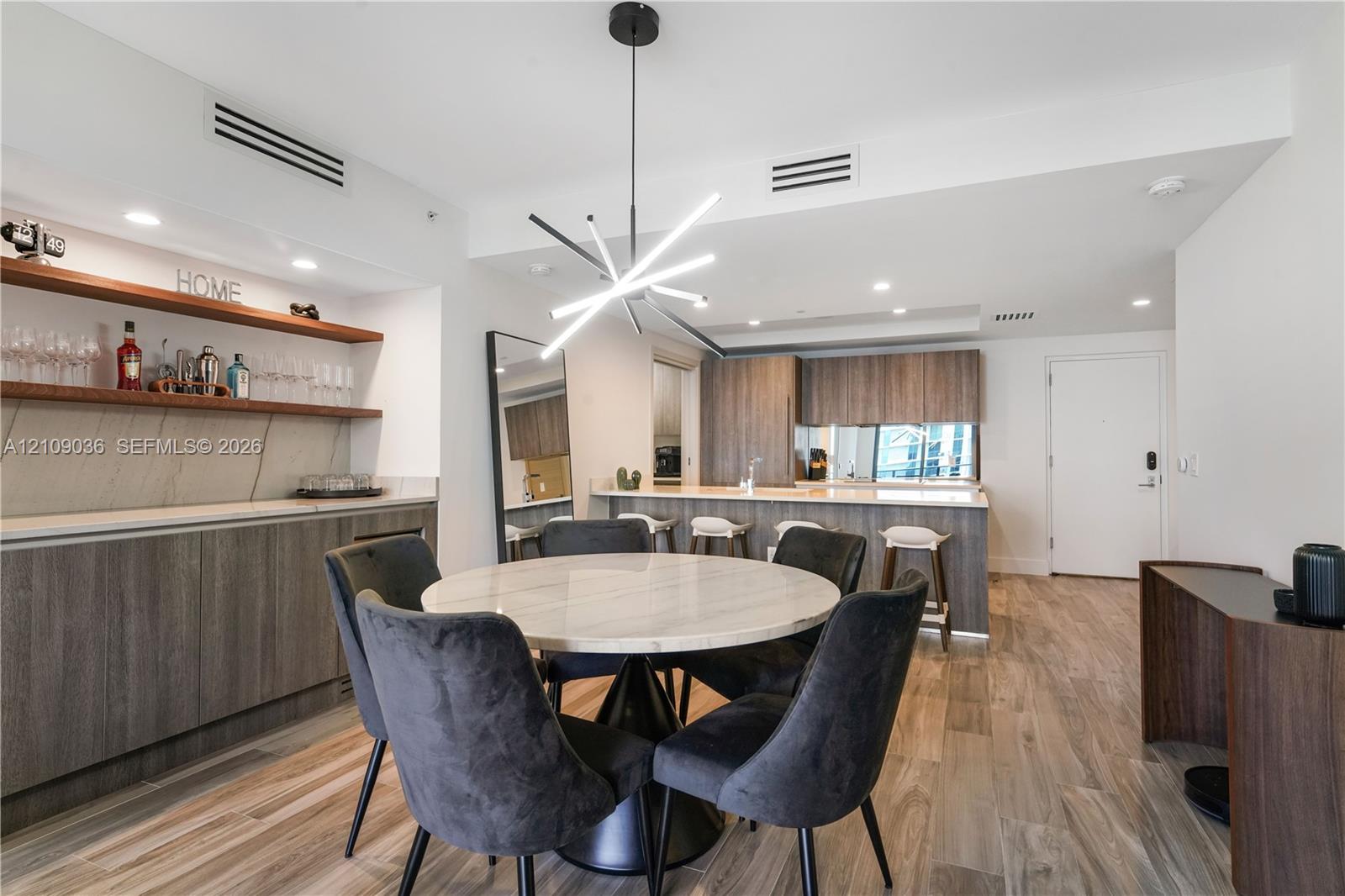 Custom wet bar featuring wood-finish cabinetry, stone-style backsplash, and floating wood shelving, complemented by wood-finish flooring, a modern geometric light fixture, and recessed lighting