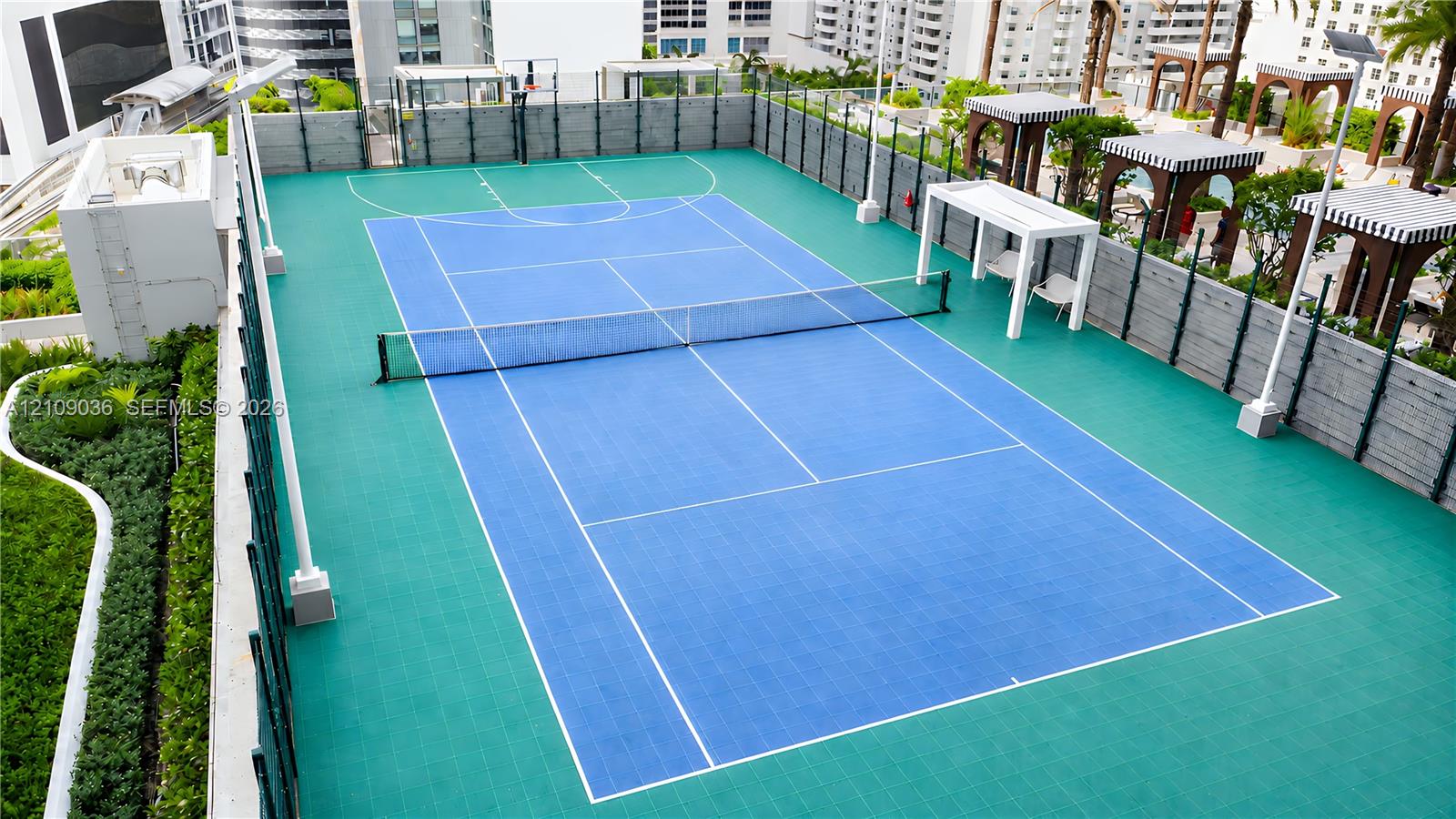 Rooftop multi-use athletic court featuring a synthetic surface, integrated basketball hoop, tennis net, and surrounding perimeter fencing with shade structures