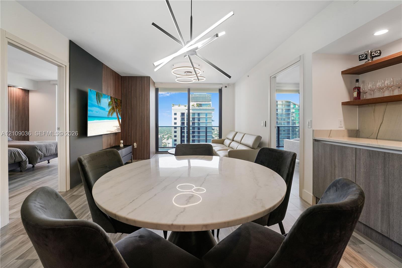Modern living space featuring floor-to-ceiling windows, wood-panel accent wall, built-in wet bar with shelving, integrated recessed lighting, and a linear architectural chandelier